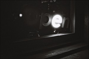 A black and white photograph of a neon sign in a window, partially reading "open." The illuminated "pen" part of the sign contrasts sharply with the dark surroundings, making it stand out. In this scene of errance, the light glows softly, casting a gentle reflection on the window's surface.